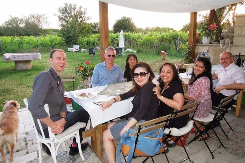 A group of people seated at an outdoor table with a white tablecloth, surrounded by greenery and a stone bench. 3