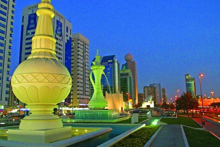 Un paisaje urbano al anochecer con edificios iluminados y dos esculturas destacadas en primer plano. 1
