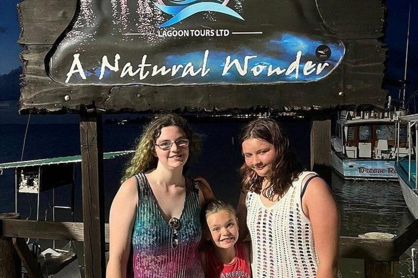 A sign with 'A Natural Wonder' and 'LAGOON TOURS LTD' text, a wooden dock, and a boat in the background. 2