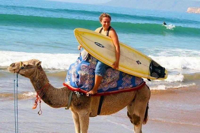 A person riding a camel with a surfboard on a beach. 1