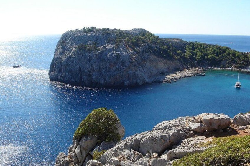 A rocky coastline with a small island, clear blue sea, and a sailboat. 3