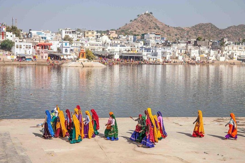 A group of women in colorful traditional attire sitting by a river, with a town and hills in the background. 4
