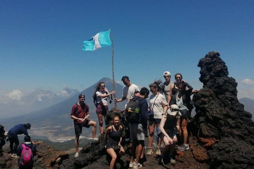 Un grupo de personas en la cima de una montaña con una bandera, formaciones rocosas volcánicas y una cordillera distante. 4
