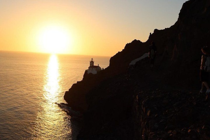 Un coucher de soleil sur une côte rocheuse, avec un phare et des personnes qui se promènent. 1