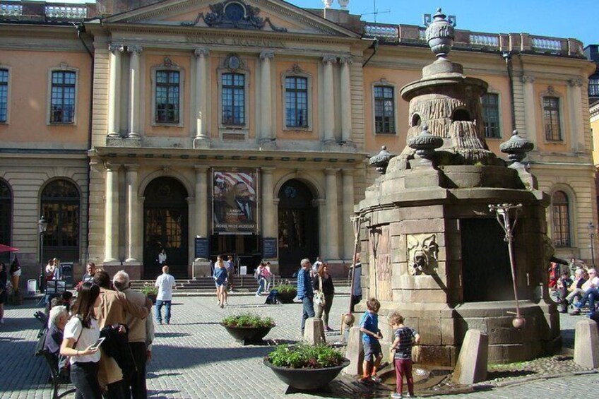 A public square with a fountain, people walking, and a historic building with columns. 5