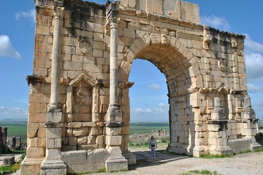An ancient stone archway with columns, set against a clear sky and green landscape. 3
