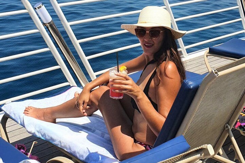 A person relaxing on a sun lounger on a boat, holding a drink with a straw. 1