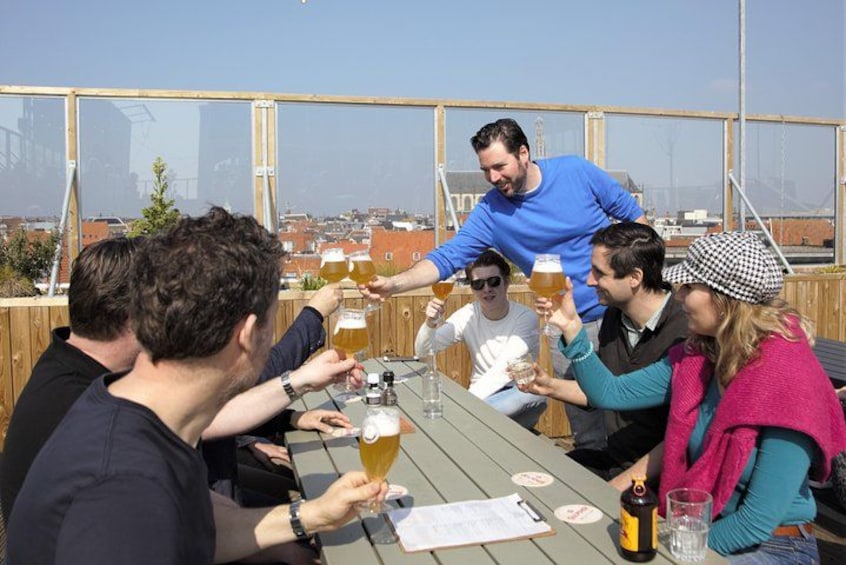 Un groupe de personnes trinquant avec des verres de bière autour d’une table en bois, sur un toit-terrasse. 1