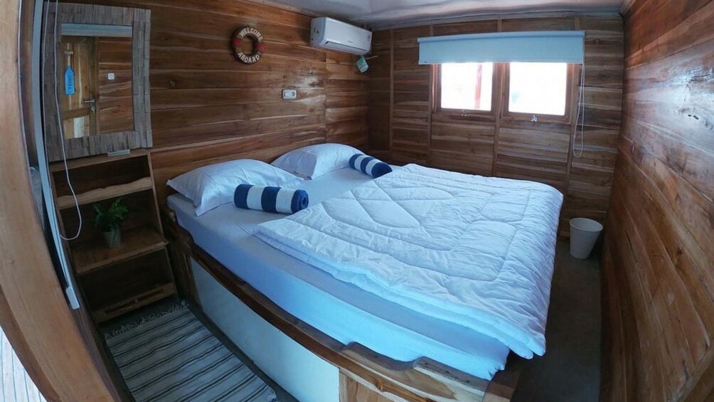 A wooden cabin bedroom with a bed, a small shelf with a plant, a welcome sign, and a window with blinds. 5