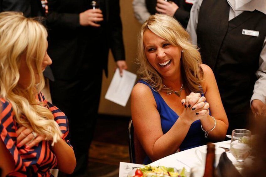 A woman in a blue dress seated at a table with a plate of food, a man in a suit standing behind her, and another woman in a patterned dress. 3