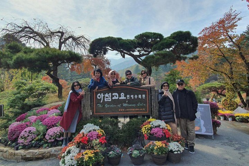 A group of people standing in front of a sign with Korean text, surrounded by various potted plants and trees. 4