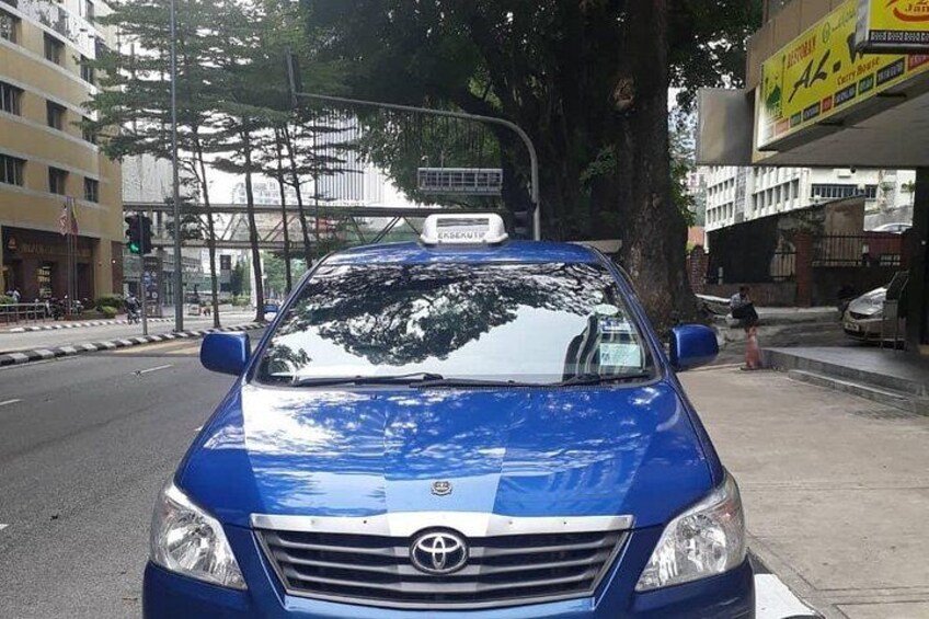 A blue car parked on a city street with buildings and a tree in the background. 2