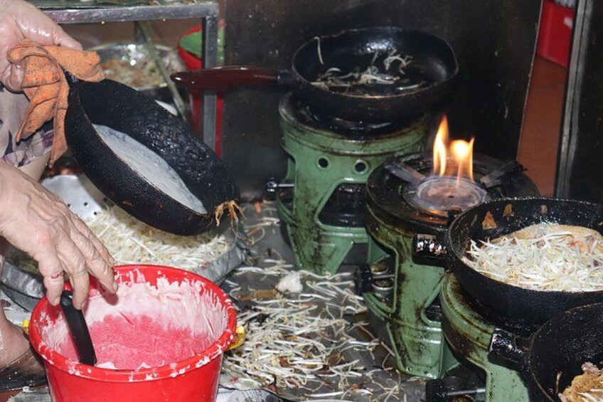 A cooking scene with a person handling a large black pan, a red bucket, and a green stove with a visible flame. 5