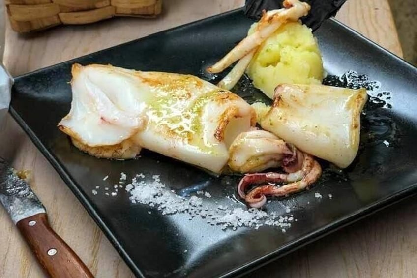 Een zwart bord met gegrilde vis en inktvis, inclusief een mes en een houten snijplank. 3