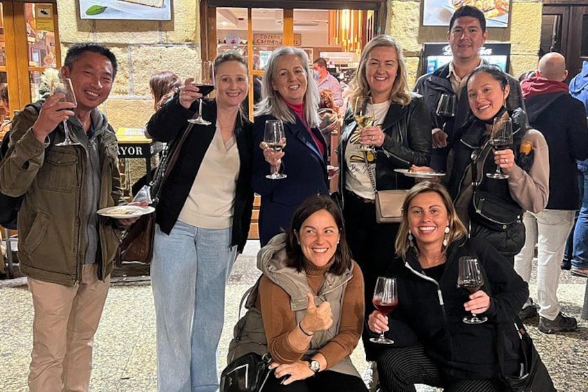A group of people holding wine glasses, standing and sitting in an outdoor setting with a stone wall and a sign that reads 'Café'. 1