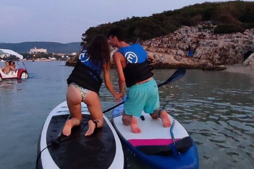 Two people on a paddleboard in calm waters, with rocky terrain and a building in the background. 1