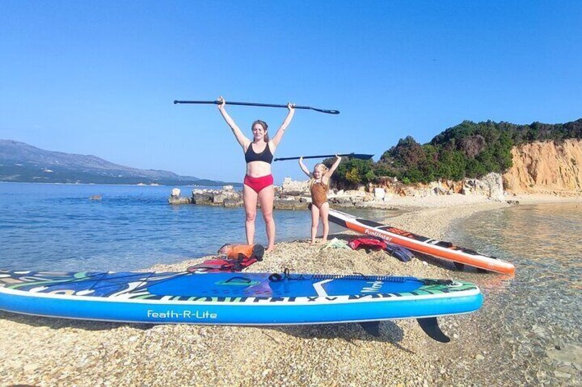 Two people standing on a paddleboard with a clear blue sky and a rocky shoreline in the background. 2