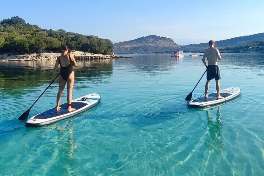 Two people paddleboarding on a calm lake with clear water and a hilly landscape in the background. 1