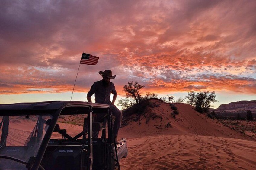 Una persona con sombrero de vaquero sentada en un vehículo en un desierto con dunas de arena y un atardecer. 3