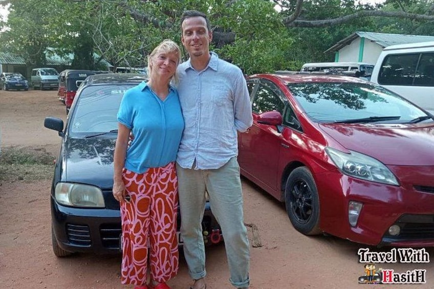 Two people standing in front of a red car and a black car in a parking lot with trees and buildings in the background. 3