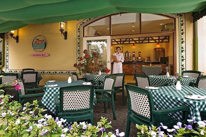 An outdoor restaurant with checkered tables and chairs, a chef at the counter, and a garden with flowers. 2