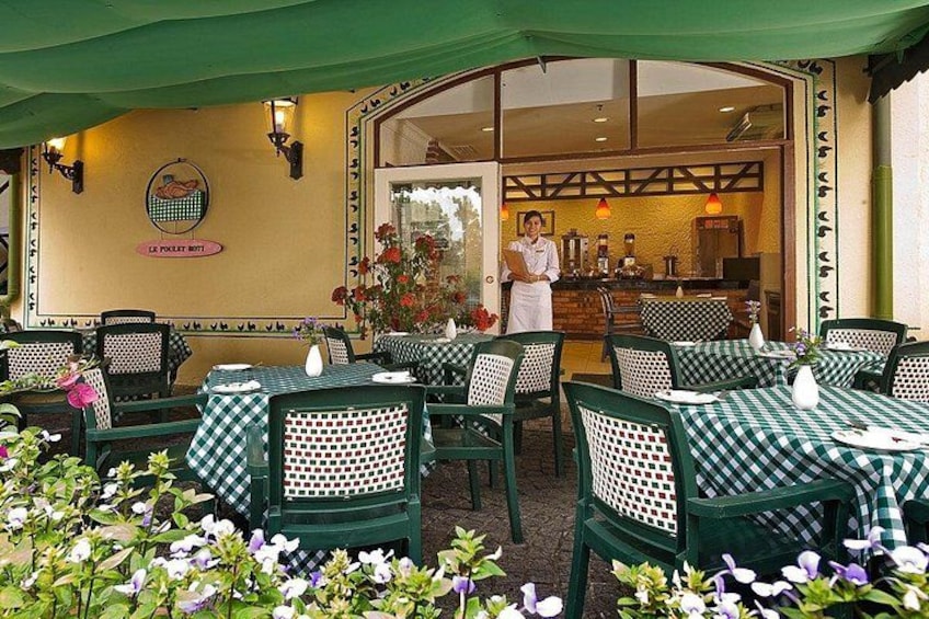An outdoor restaurant with checkered tables and chairs, a chef at the counter, and a garden with flowers. 2