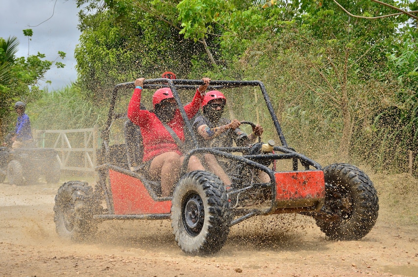 A red off-road vehicle with two people in red helmets driving through a dusty area. 1