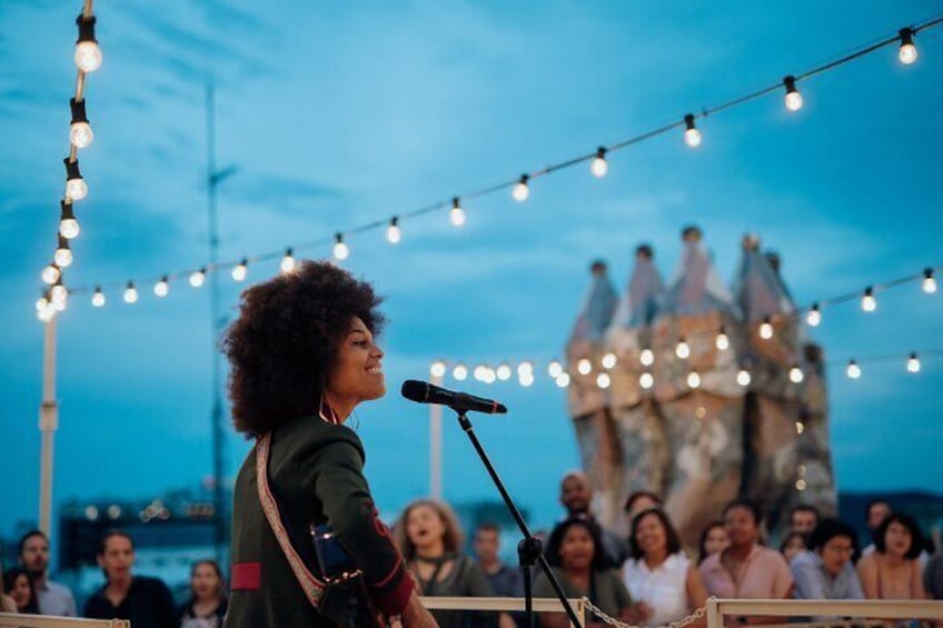 A person performing on stage with a microphone, audience in the background, and string lights overhead. 4
