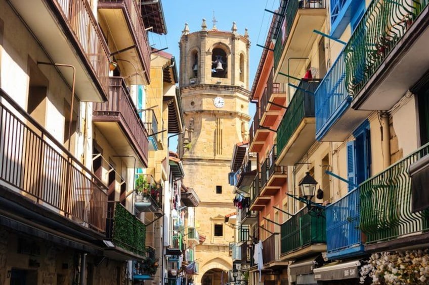 A narrow street with buildings on either side, a bell tower, and a clock. 5