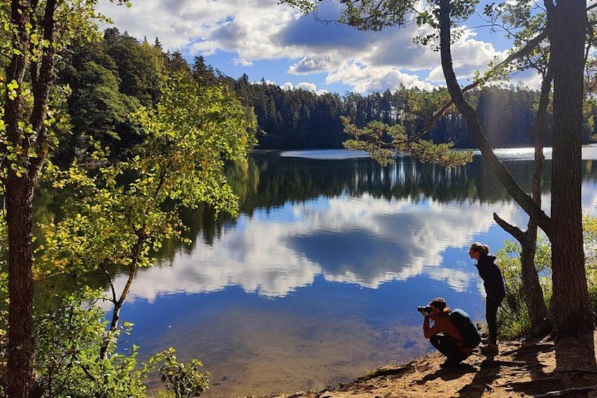 A lake surrounded by trees, a person taking a photo, and another person sitting on the shore. 1