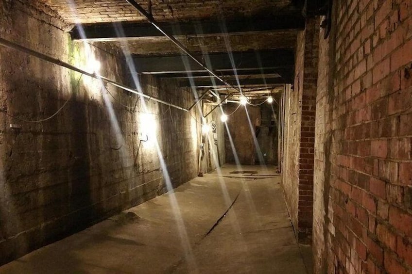 A dimly lit underground passageway with brick walls and concrete floor, illuminated by overhead lights. 2