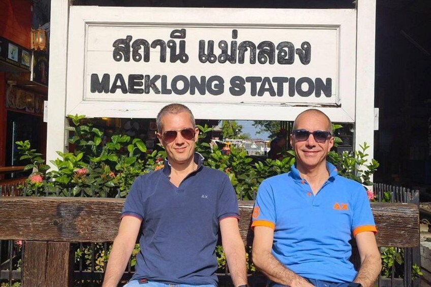 Två män sitter på en bänk framför en skylt där det står ”MAEKLONG STATION”. 5