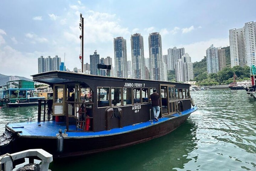 A ferry named 'Jumbo Ferry 7' with a blue and brown color scheme, docked at a harbor with a city skyline in the background. 1