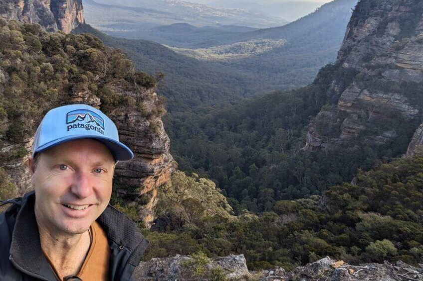 A person wearing a Patagonia cap standing on a rocky outcrop overlooking a valley with dense forest and cliffs. 3
