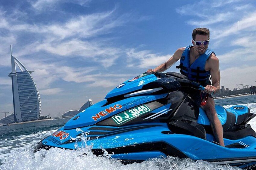 A person riding a blue jet ski with the text 'NEMO' and 'DJ 3845' on it, in a body of water with a sail-shaped building in the background. 4