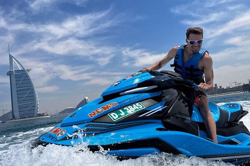 A person riding a blue jet ski with 'NEMO' and 'DJ 3845' on the side, in a body of water with a sail-shaped building in the background. 4