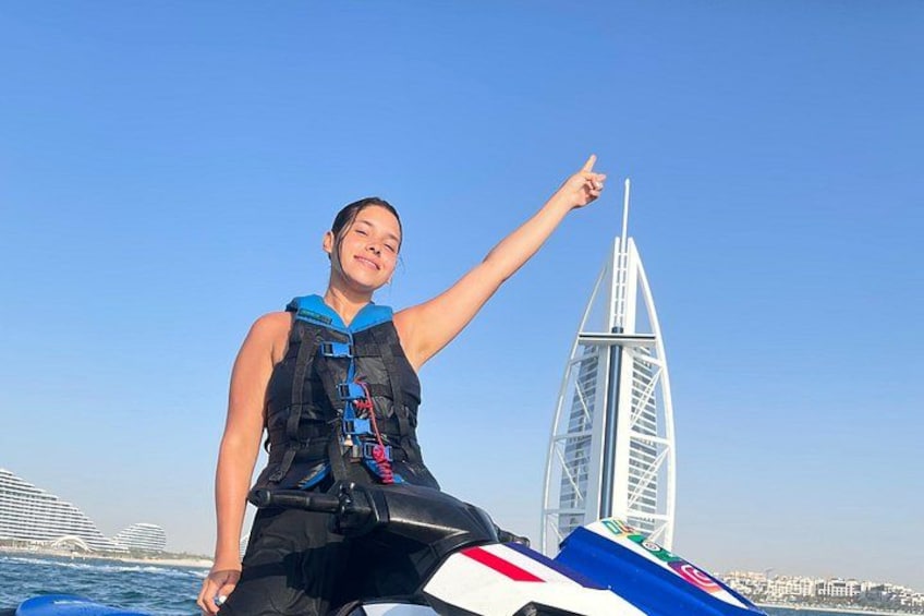 A person on a jet ski with a life jacket, pointing towards a distinctive sail-shaped building in the background. 3