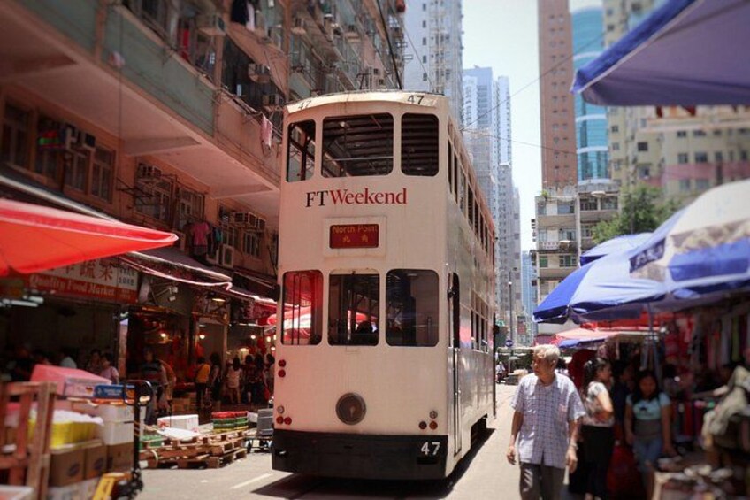 Ein Doppeldeckerbus mit der Aufschrift 'FTWeekend' auf einer belebten Straße mit Marktständen und Hochhäusern. 1