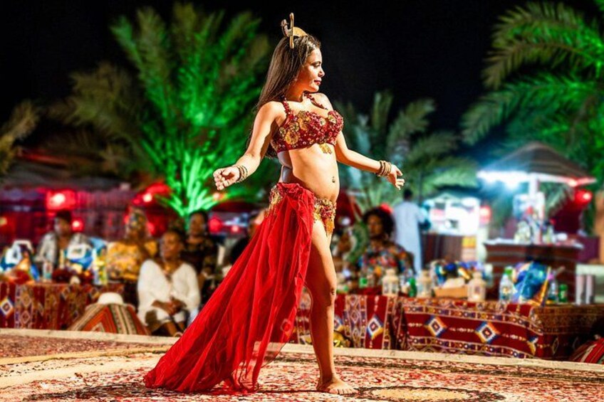 A belly dancer performing on a patterned carpet with palm trees and a crowd in the background. 2