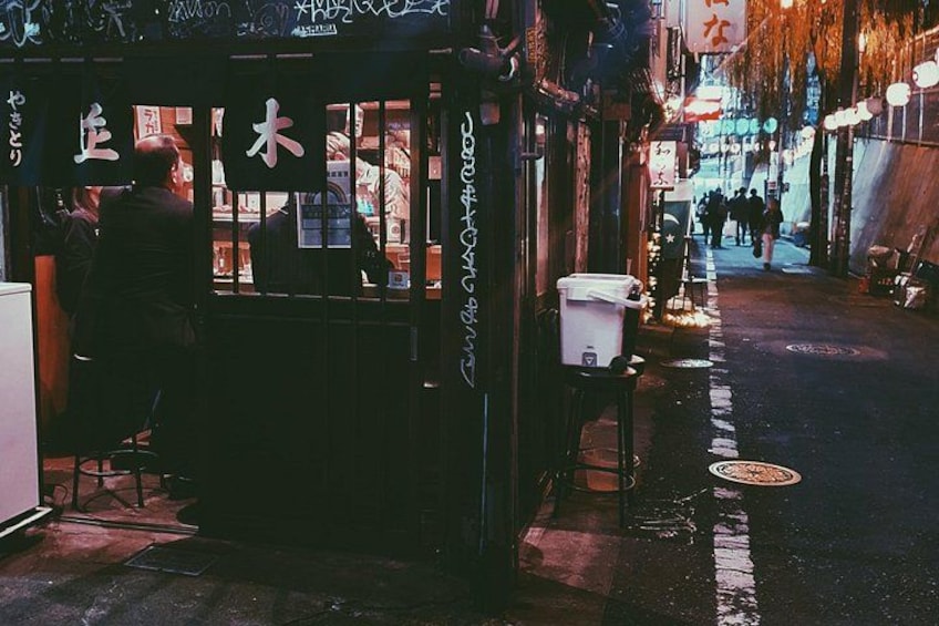 Una calle estrecha con una pequeña tienda a la izquierda, un bote de basura blanco a la derecha, y gente caminando al fondo. 3