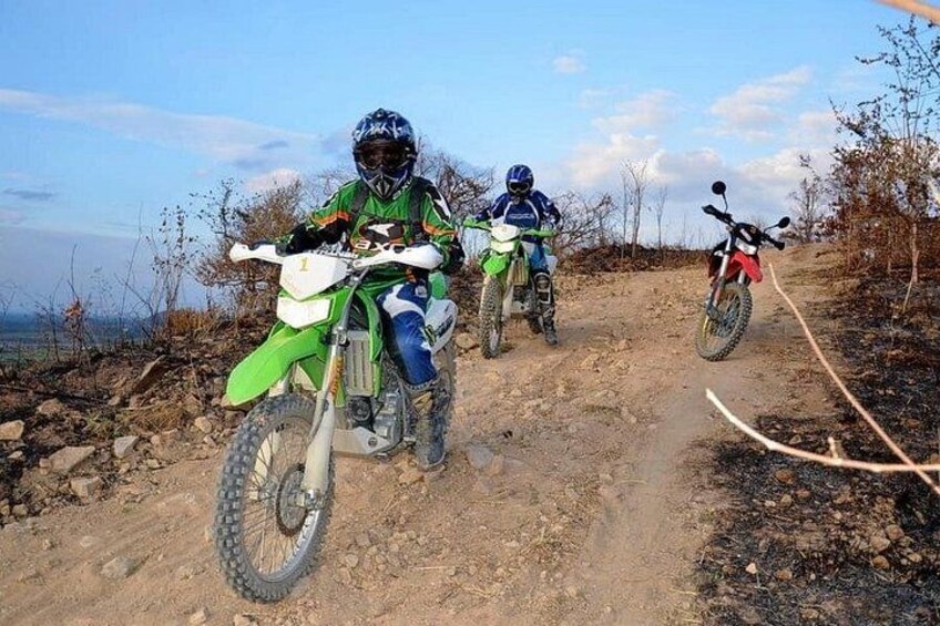 Three dirt bikes on a dirt trail with rocky terrain and sparse vegetation. 1