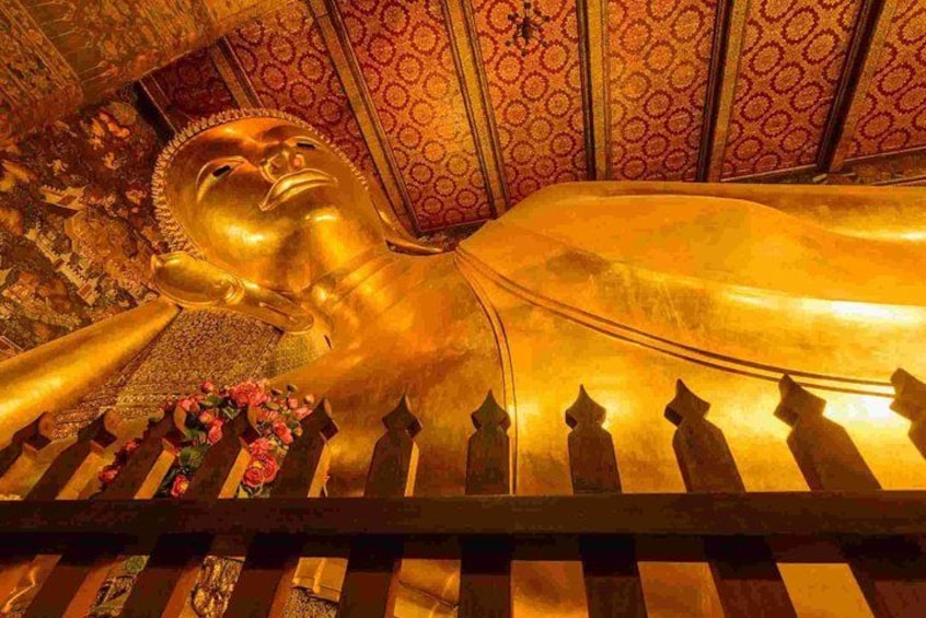En stor, gylden liggende Buddha-statue med detaljerede udskæringer, omgivet af dekorative røde vægge med mønster og et trægelænder med blomsterudsmykning. 2