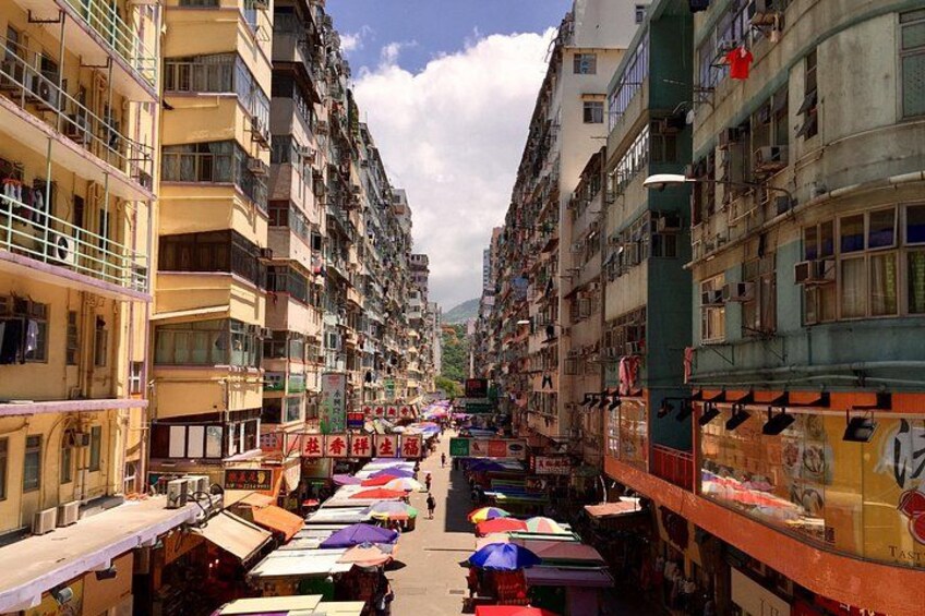 A narrow street flanked by multi-story buildings with balconies, vendors with colorful umbrellas, and a clear sky. 5