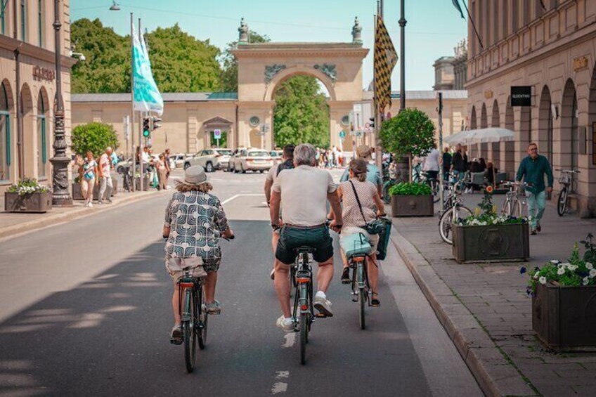 A street with people riding bicycles, a large archway, and buildings with historical architecture. 2