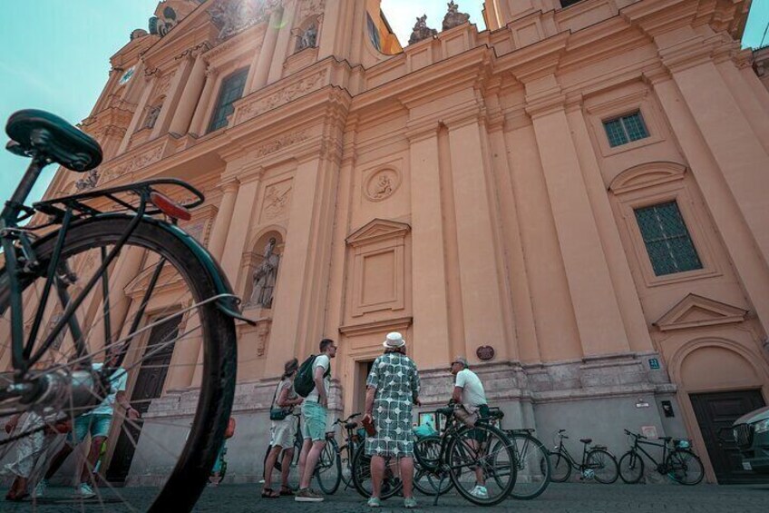 A large pink building with a clock and statues, bicycles parked in front, and people standing nearby. 3