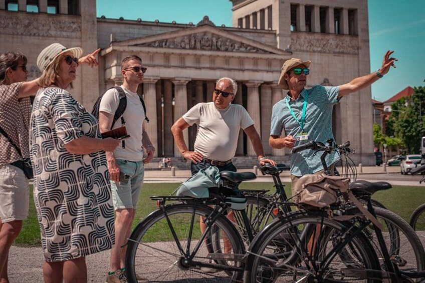 A group of people standing in front of a building with classical architecture, with bicycles parked nearby. 1