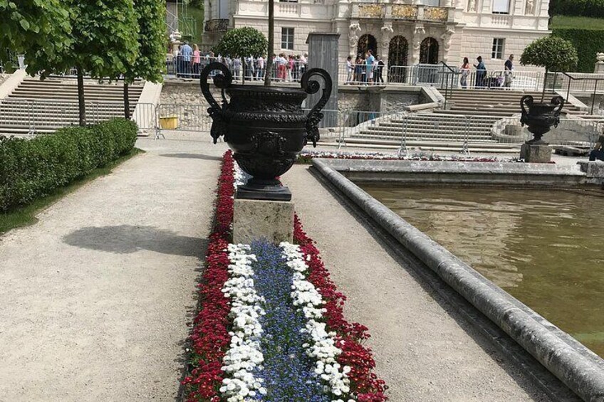 A pathway lined with flowers, a large black urn, and a fountain. 4