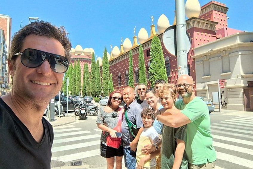 A group of people posing for a photo in an urban setting with distinctive architecture, including a building with large spherical domes and a street with parked cars. 5