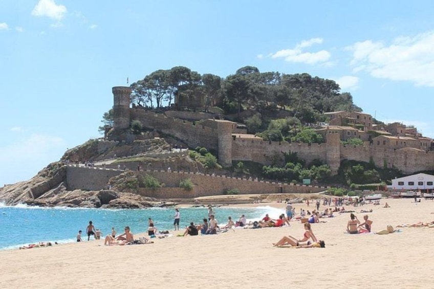 Une plage où des gens se prélassent au soleil et se baignent, un château perché sur une colline et un ciel bleu azur. 2
