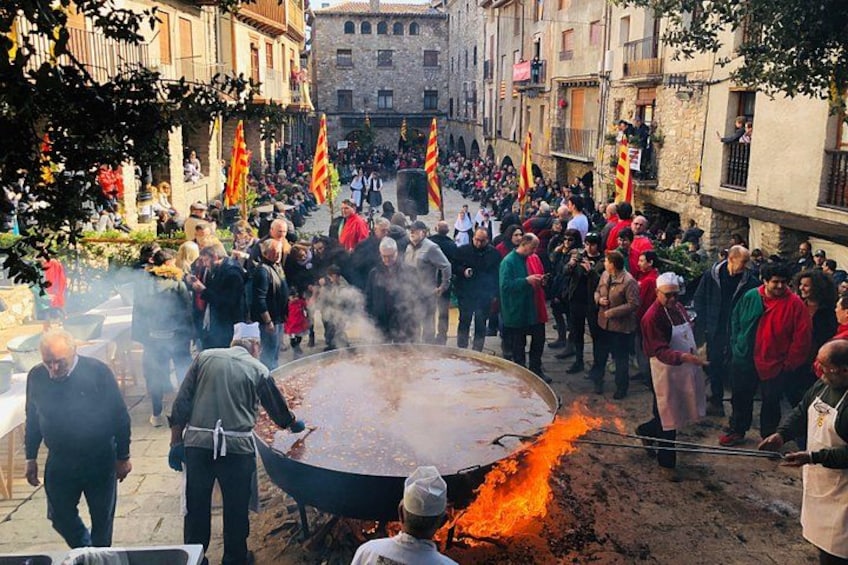 A large outdoor cooking event with a circular grill, people gathered around, and flags. 4